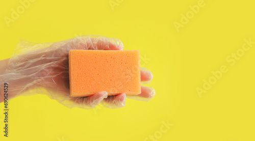 Orange dishwashing sponge in a woman's hand on a yellow background. A hand in a transparent disposable glove holds a sponge for wet cleaning. Professional cleaning. Copy space