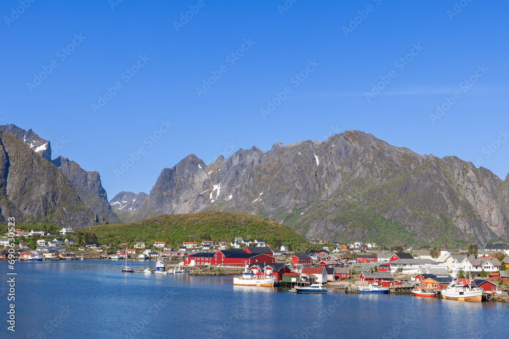 Panoramic view of Reine featuring classic red Rorbu houses and moored fishing boats at the docks, set against majestic mountains with snow-capped peaks in the Lofoten Islands, Norway