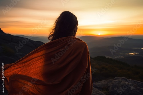 Person wrapped in an orange blanket watching the sunrise from a mountaintop, symbolizing contemplation and peace.