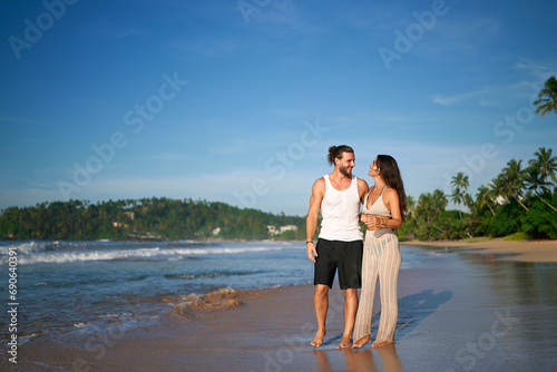 Romantic couple strolls on beach, holding hands by the sea. Man, woman enjoy honeymoon at tropical coast. Casual beachwear, sunset walk, love, vacation. Smiling duo in relaxed seaside promenade.