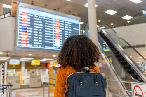 Black woman looks at the flight schedule on a digital monitor in an airport to check the gate and time to board the plane to travel or study abroad.