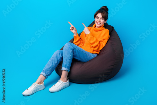 Photo of positive good mood woman dressed sweater sitting bean bag pointing two fingers empty space isolated blue color background