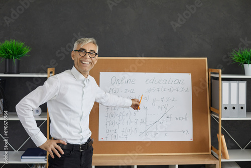 Senior smiling professional math teacher stands and points to a board with mathematical equations. Male lecturer with glasses explains mathematical formulas to students. Online education concept.