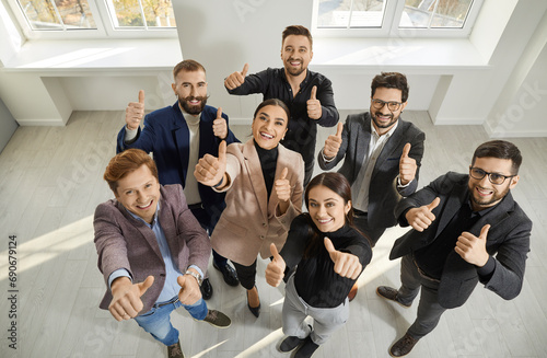 Team of happy, satisfied business people giving thumbs up all together. Joyful young men and women standing in office, looking up, smiling and doing like gestures. Shot from above. Success concept
