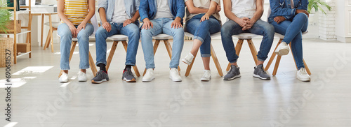 Diverse group of different young people wearing casual shirts, modern blue jeans and white and gray sneakers sitting on row of chairs in office workplace. Cropped low section shot of human legs photo