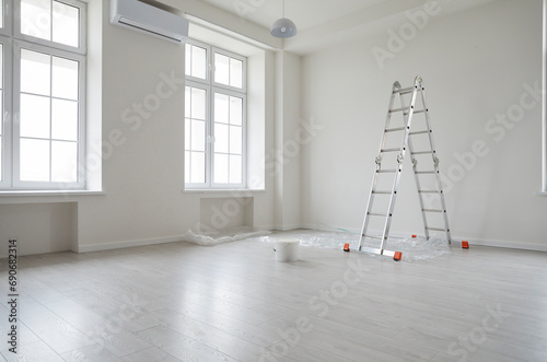Empty unfurnished room with ladder and paint bucket. Unfinished living room interior with white walls, floor and windows. Renovation. Repairs in apartment. Empty interior space