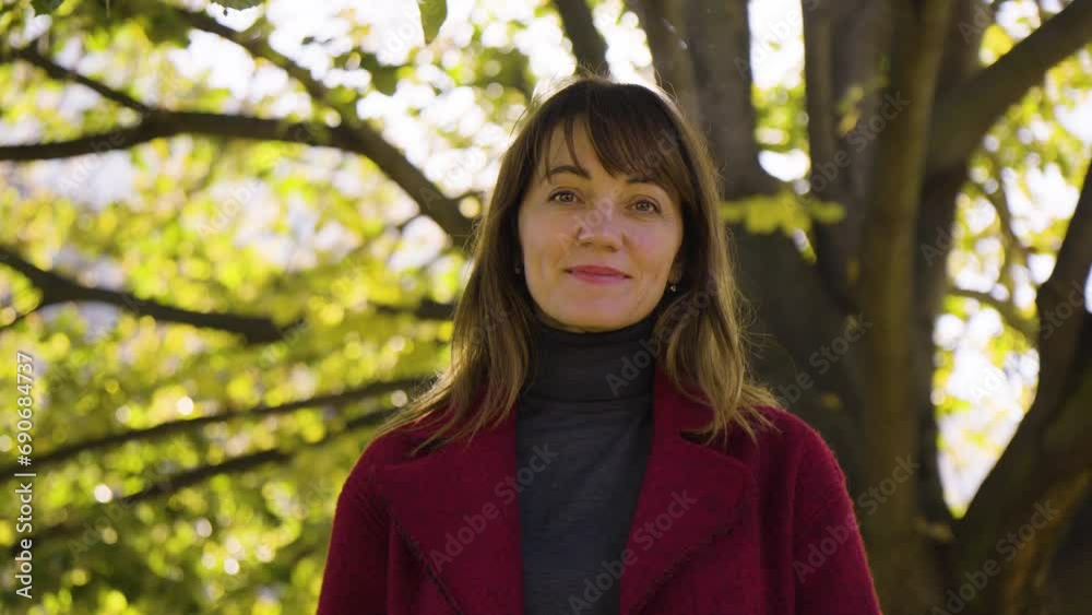 An attractive middle-aged Caucasian woman waves at the camera with a smile in a park - closeup from below - behind her, sun shines through a tree crown An attractive middle-aged Caucasian woman waves at the camera with a smile in a park - closeup from below - behind her, sun shines through a tree crown