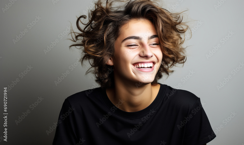 Joyful young person with short curly hair wearing a black t shirt, laughing and looking at the camera against a light grey background Joyful young person with short curly hair wearing a black t shirt, laughing and looking at the camera against a light grey background