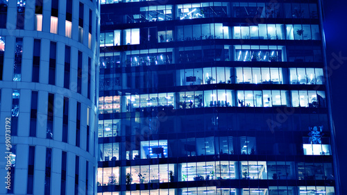 Fragment of the glass facade of a modern corporate building at night. Big glowing windows in modern office buildings at night, in rows of windows light shines. Blue graphic filter.