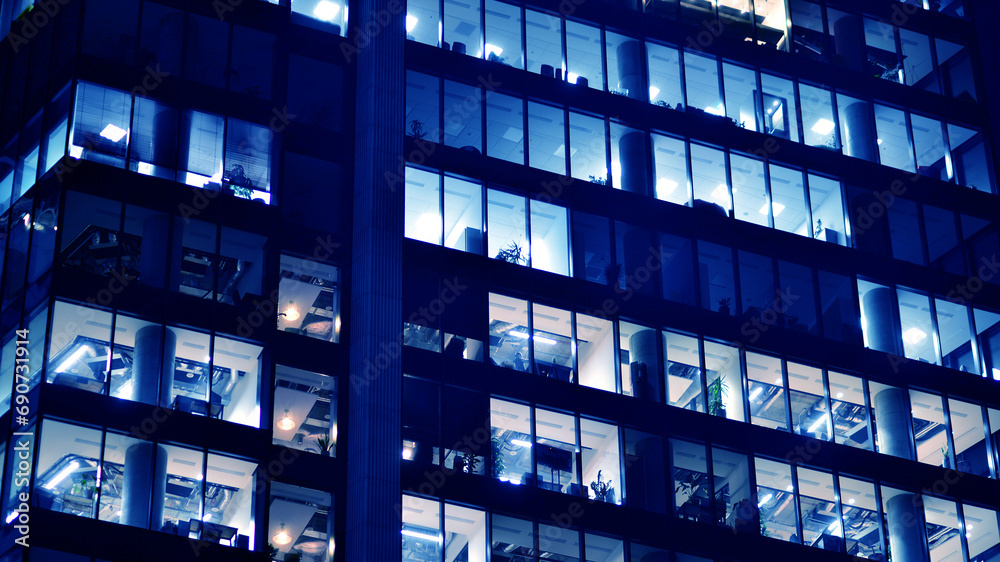 Fragment of the glass facade of a modern corporate building at night. Big glowing windows in modern office buildings at night, in rows of windows light shines. Blue graphic filter.