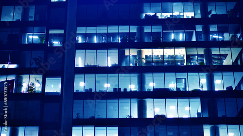 Fragment of the glass facade of a modern corporate building at night. Big glowing windows in modern office buildings at night, in rows of windows light shines. Blue graphic filter.