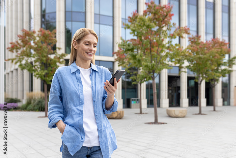 Mature woman with phone in hands walking in the city, a businesswoman in blue shirt holding smartphone in hands, reading online social networks, blonde smiling satisfaction is using an application.