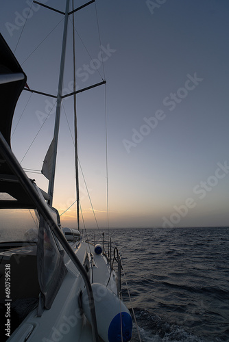 sailing ship glides gracefully on the sea as the sun sets. Its sails are being lowered, creating a serene atmosphere. The vibrant sunset colors reflect on the calm waters.