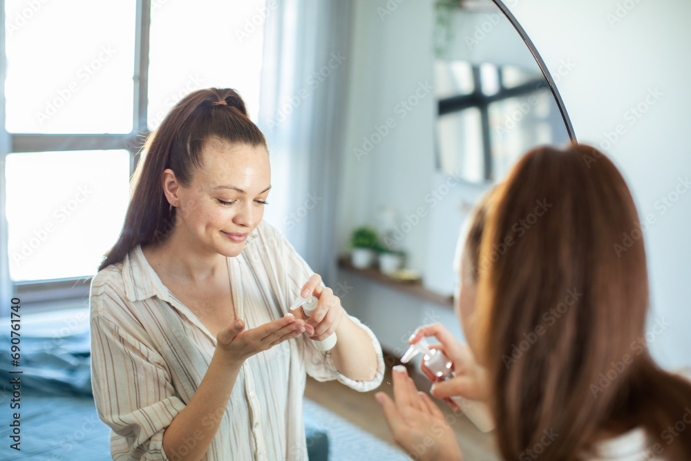 Young woman looking at mirror while touching her face applying moisturizer Young woman looking at mirror while touching her face applying moisturizer