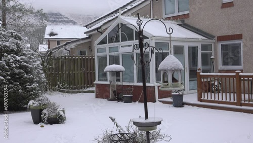 Small garden birds eating seeds from a snow topped garden feeder. In the background is a snow-covered conservatory and in the distance are snow-capped hills. photo