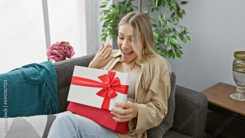 Cheerful young blonde woman, comfortably sitting on the cozy sofa at home, joyfully surprised by the beautiful ribbon gift box, utterly happy and wearing a contagious smile on her face! Cheerful young blonde woman, comfortably sitting on the cozy sofa at home, joyfully surprised by the beautiful ribbon gift box, utterly happy and wearing a contagious smile on her face!