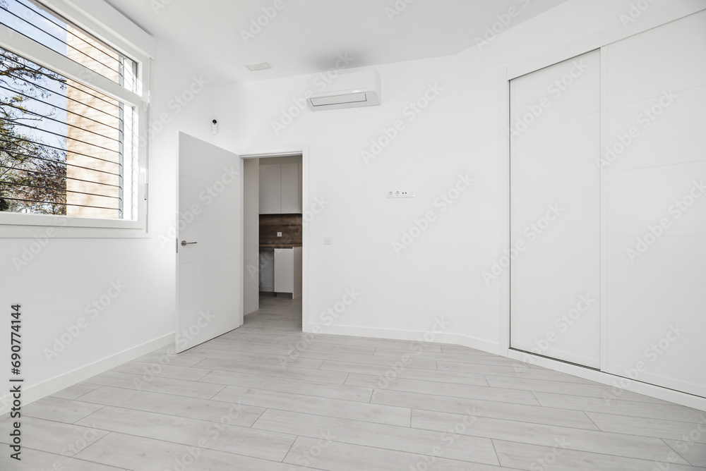 An empty room with a bay window in a loft-style house with a two-section built-in wardrobe with white wooden sliding doors, access to a room with a kitchen and light wooden floors