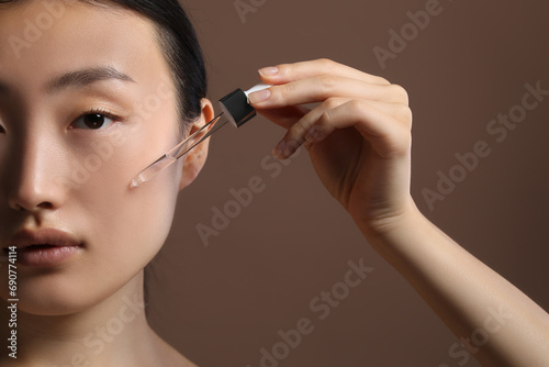Beautiful young woman applying cosmetic serum onto her face on brown background  closeup