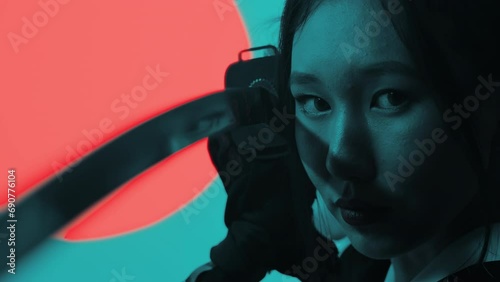 Cinematic close-up of a young asian woman in an epic militant pose holding a katana near her face in the studio with a turquoise blue green background and red sun photo