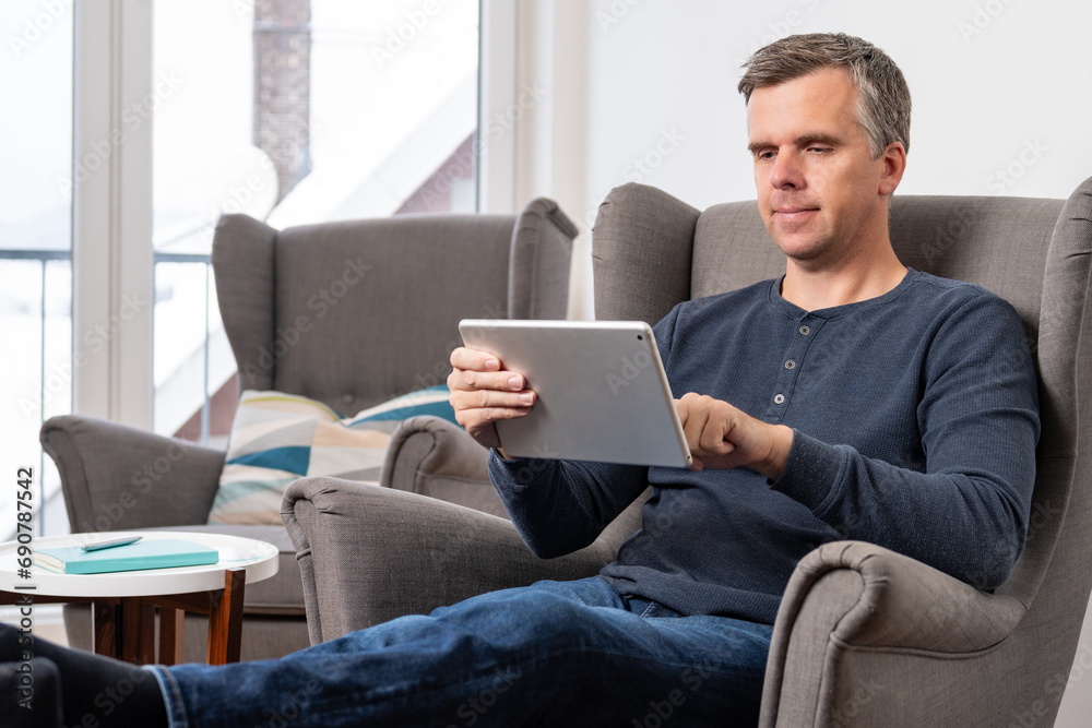 handsome 40s single man sits in an comfy  grey armchair and uses tablet in landscape mode
