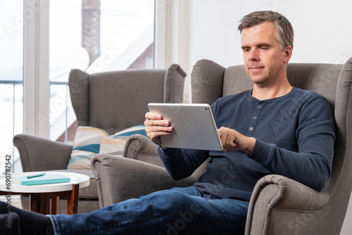 handsome 40s single man sits in an comfy  grey armchair and uses tablet in landscape mode

