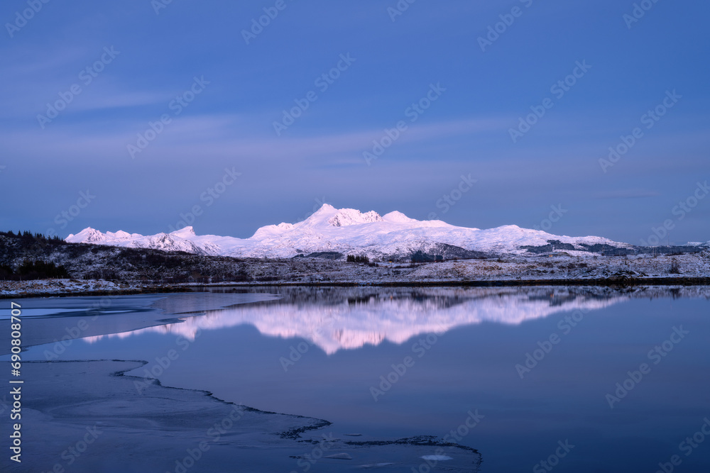 Naklejka premium Snowy mountains, sea bay, reflection in water at sunset in winter. Lofoten islands, Norway. Colorful Landscape with rocks in snow, blue sky at dusk. Seashore at dusk. Nature background. Scenery