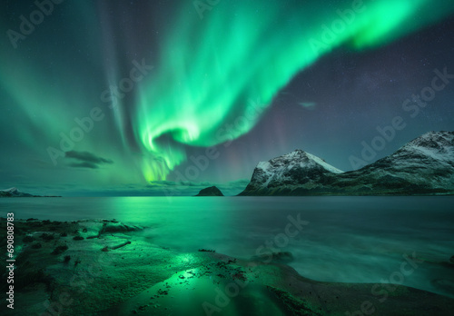 Aurora borealis over the sea, snowy mountains at starry winter night. Northern Lights in Lofoten islands, Norway. Sky with polar lights. Landscape with aurora, rocky beach, sky, reflection in water