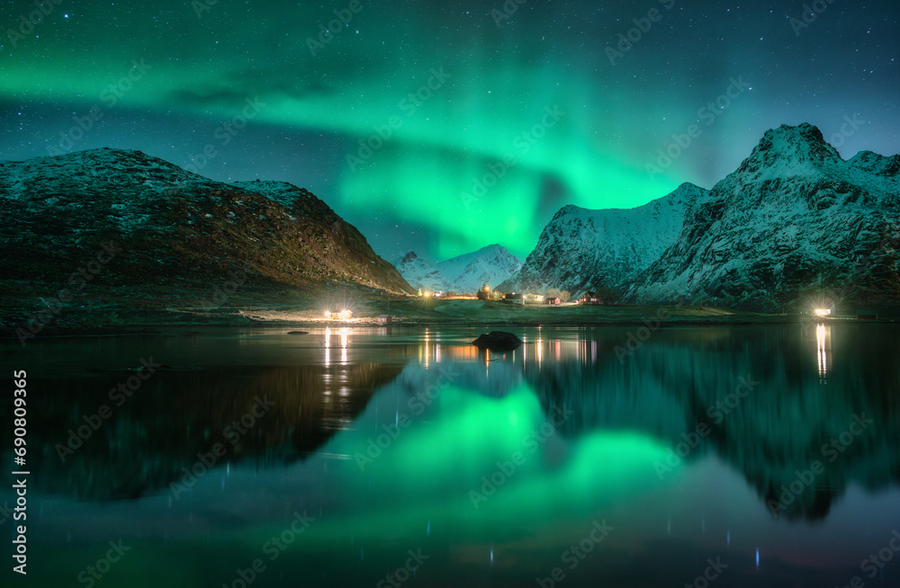 Aurora borealis, snowy mountains, sea, fjord, reflection in water, street lights at starry winter night. Lofoten, Norway. Northern lights. Landscape with polar lights, snowy rocks, sky with stars - obrazy, fototapety, plakaty 