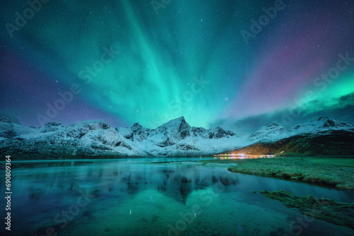 Northern lights over the snowy mountains, frozen sea, reflection in water at winter night in Lofoten, Norway. Aurora borealis and snowy rocks. Landscape with polar lights, starry sky and fjord. Nature