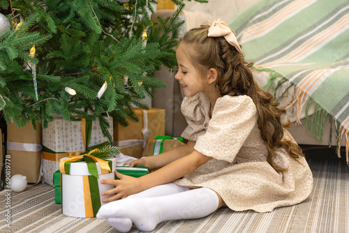Christmas spirit. Happy little smiling girl opening gift box sitting on the floor near the Christmas tree. Happy girl unwrapping christmas present looking in box. Holidays and celebrations concept