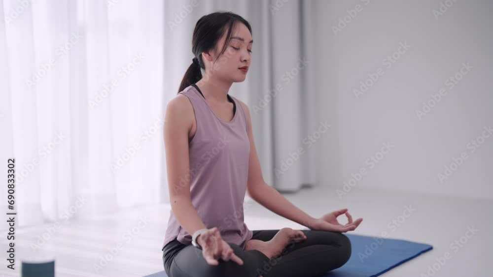 Asian woman in workout attire practices meditation on a yoga mat in her home's sitting room. Perfect for showcasing relaxation and mindfulness in a domestic setting. Asian woman in workout attire practices meditation on a yoga mat in her home's sitting room. Perfect for showcasing relaxation and mindfulness in a domestic setting.