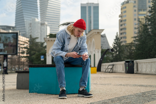 Pensive hipster man sitting in urban street with smartphone, reading, messaging, searching information, posting photos in social networks. Slim guy looks like blogger, tourist, international student