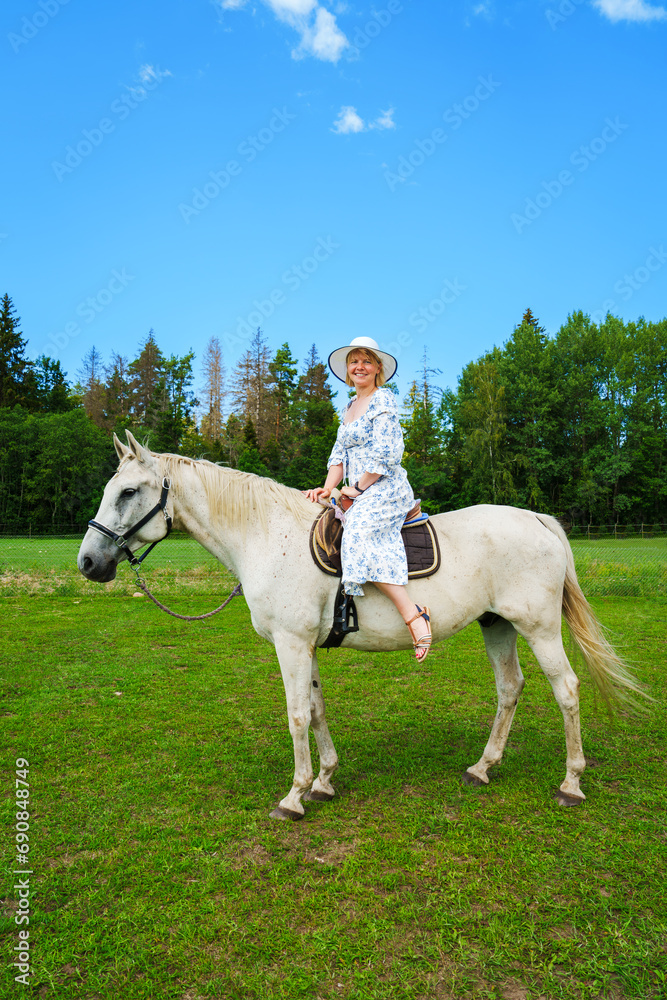 Obraz premium A stunning young woman with flowing hair enjoying a leisurely ride on a graceful white horse amidst picturesque countryside on a radiant, sunlit summer day.