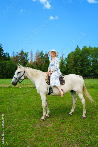 A stunning young woman with flowing hair enjoying a leisurely ride on a graceful white horse amidst picturesque countryside on a radiant, sunlit summer day. © Ilja
