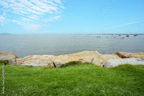 sea ocean to marine and blue sky with many small wooden fishing boats and jetty harbor to green grass lawn and rocks foreground to summer with copy space at Bang Phra Public Park Chon Buri in Thailand