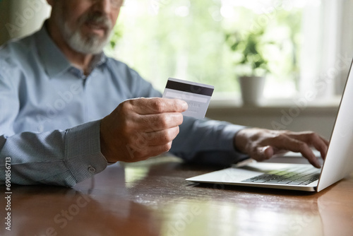 Aged ebank client. Close up view of older man retiree holding credit plastic card paying bills on laptop online buying goods using ecommerce web app. Senior male customer make internet payment via pc photo
