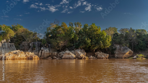 Sheer limestone cliffs rise on the riverbank. Ripples and the reflection on the shiny red-brown water. Green vegetation against a background of blue sky and clouds. Madagascar. Manambolo River