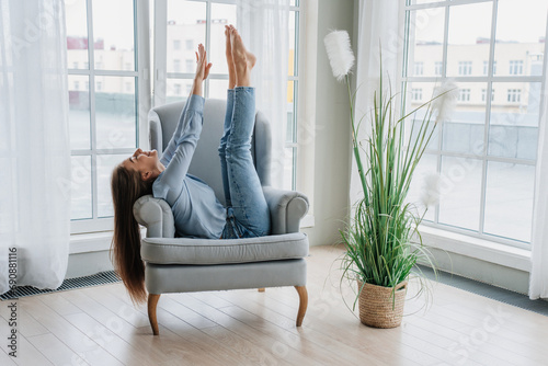 Relaxed caucasian girl in jeans sitting in cozy chair having fun rising hands and legs, doing nothing at home, reboot, weekend. Caress student enjoying vacations.