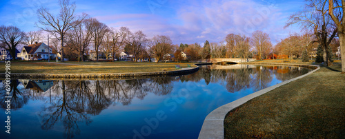 Milford Connecticut City Landscape at the Lower Lagoon Riverwalk Park in winter with curving calm river, green field, bare maple and oak trees, and traditional charming New England houses at sunset