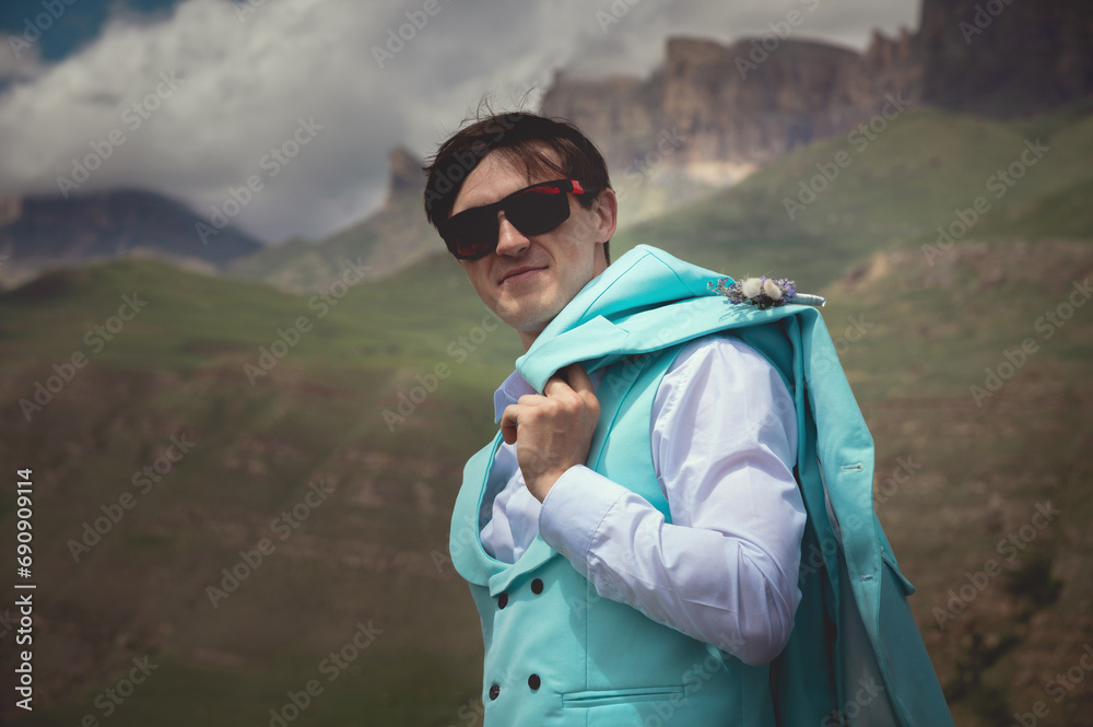 Portrait of a cheerful Caucasian guy in black glasses, holding his jacket over his shoulder and looking down at the camera, against the backdrop of rocky mountains and low clouds. confident young man