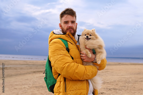 Happy young man walking with his pet Pomeranian Spitz dog on the beach, hold puppy on hands. People love animals concept.