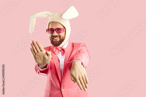 Funny extravagant man in pink suit and hat with rabbit ears dancing on pink background. Caucasian man in red glasses in formal suit and humorous hat makes funny movements looking at camera. Banner. photo