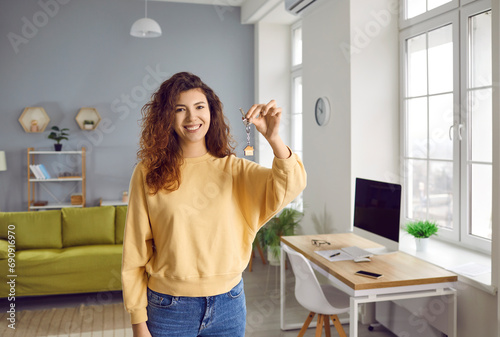 Portrait of a young smiling pretty curly redhead woman looking at the camera with the keys in her hands standing in the living room at home. Happy joyful new homeowner enjoing real estate purchase. photo