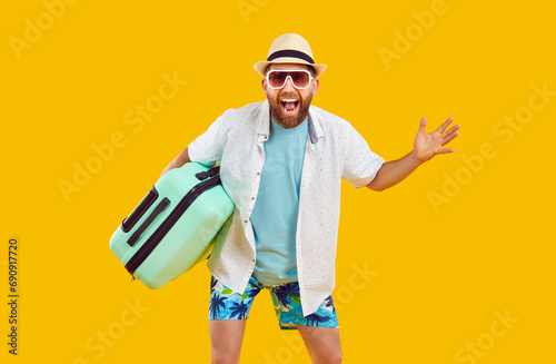 Happy male tourist with suitcase. Joyful bearded man in summer clothes, cool sunglasses and fedora sunhat holding his bag and standing isolated on yellow color background. Travel, holiday concept
