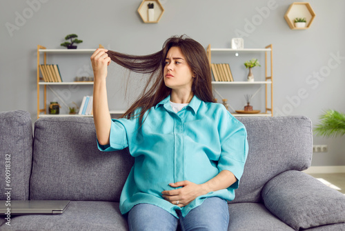 Portrait of a young brunette sad pregnant woman in casual clothes sitting on sofa in the living room at home and looking on her long hair falling out. Pregnancy and hair loss problems. photo