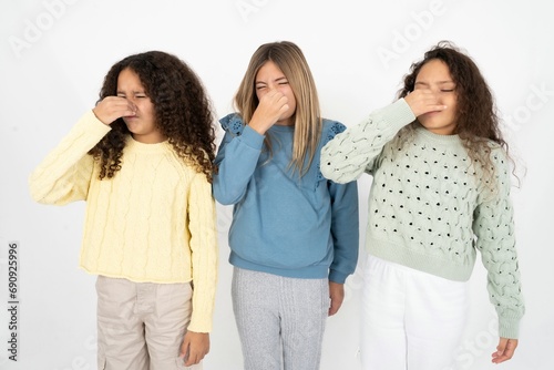 Three beautiful multiracial kid girls smelling something stinky and disgusting, intolerable smell, holding breath with fingers on nose. Bad smell