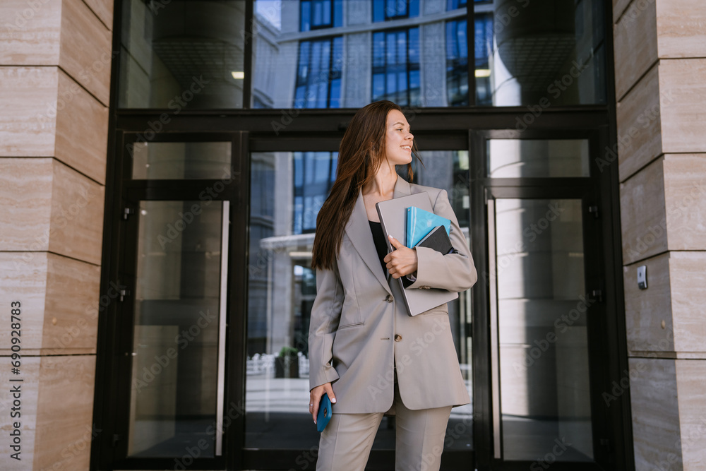 Purposeful business woman in suit standing at Entrance of bank holds laptop, notebooks, phone smiles waiting for partner. Financial people.