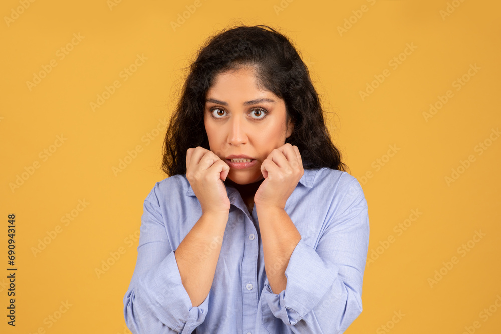 A worried young woman with curly hair wearing a blue shirt bites her nails and looks directly at the camera A worried young woman with curly hair wearing a blue shirt bites her nails and looks directly at the camera