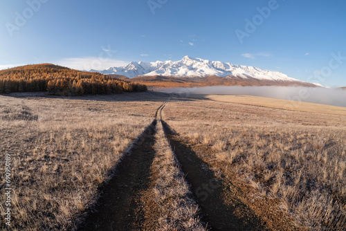 Remote dirt trail through the frozen steppe to snow mountains. Bright sunny autumn atmospheric minimalist landscape with a dirt path among the grasses in the highlands. photo