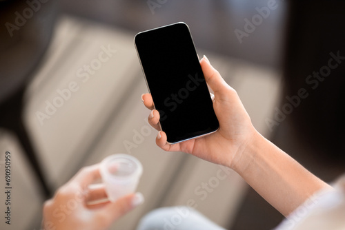 Unrecognizable woman holding smartphone blank screen and menstrual cup indoor photo